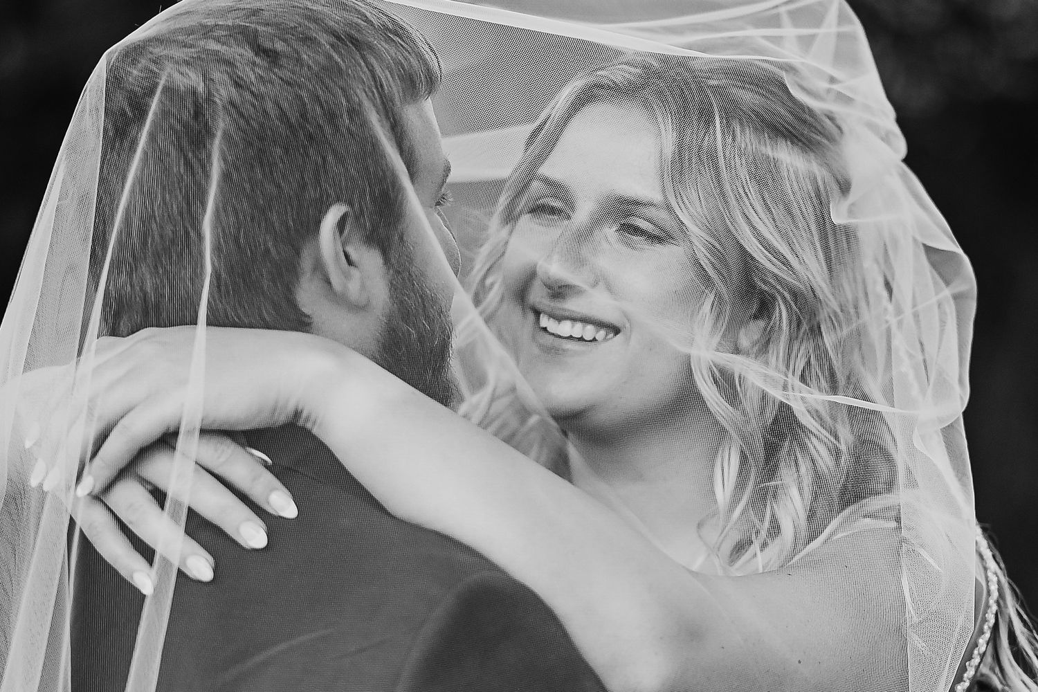 Candid black and white wedding photograph showing a happy couple embracing under a delicate veil outdoors.