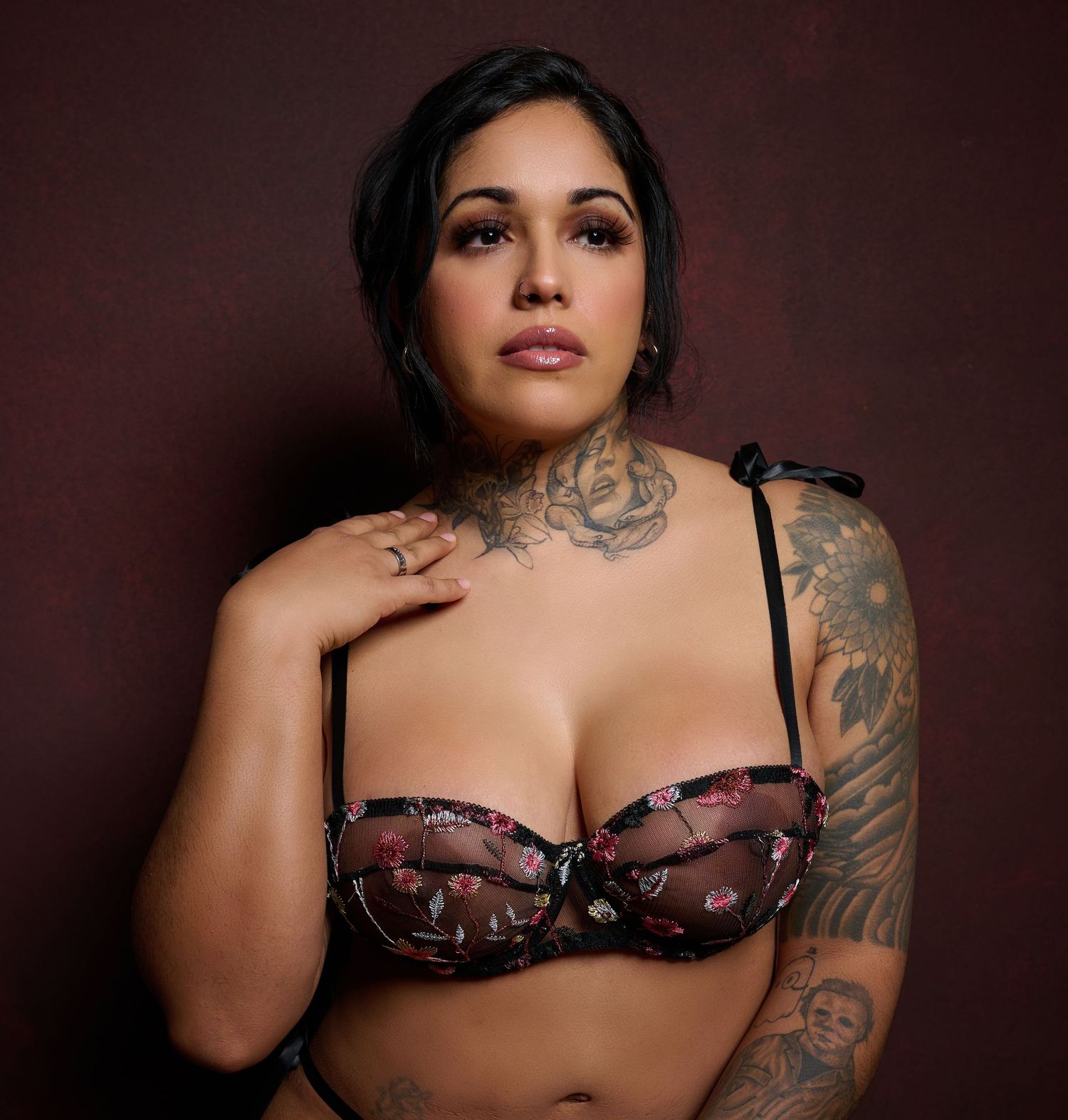 A portrait of a person wearing a burgundy bra with tattoos visible on their arm and neck against a dark background.