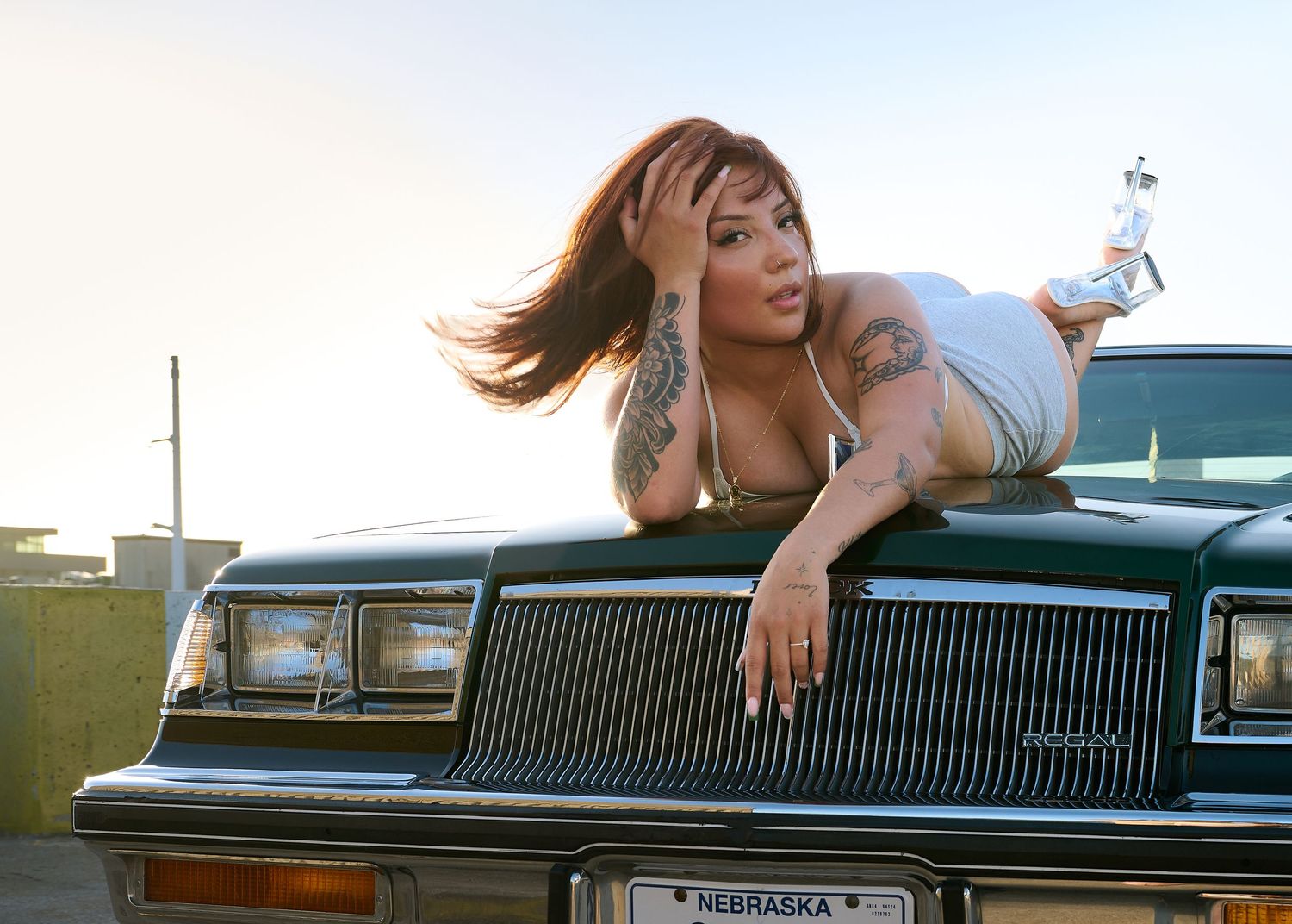 A person leans casually on the hood of a vintage Buick car during a sunset photoshoot.