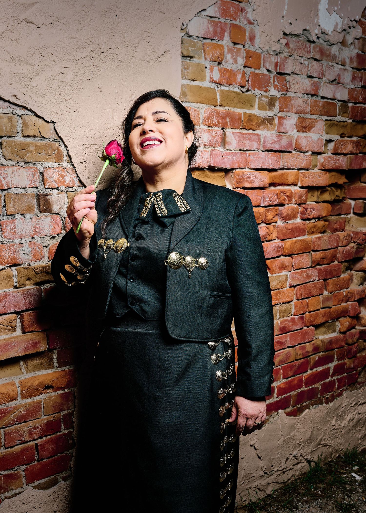 Woman in dark mariachi outfit holds a rose, smiling against a rustic brick wall.