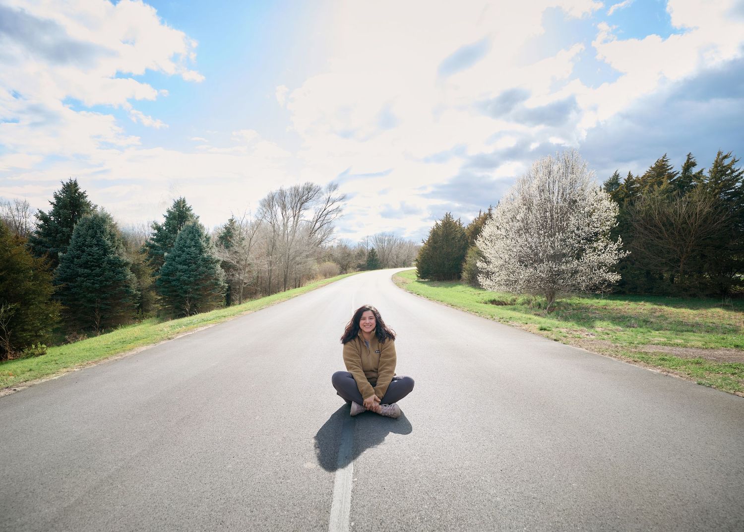 A person sitting cross-legged in the middle of an empty rural road with flowering trees in spring.