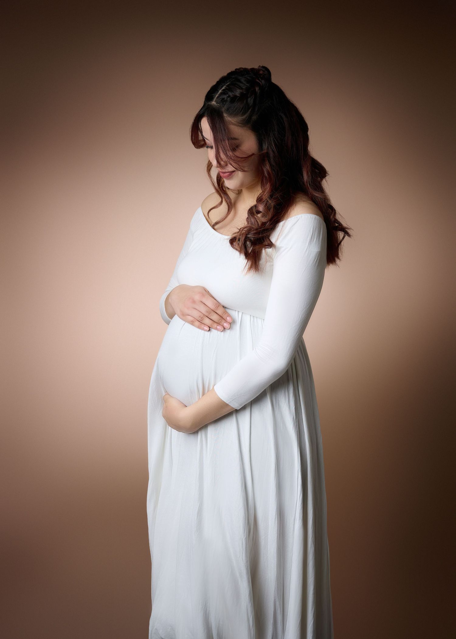 A maternity portrait featuring a woman in a flowing white dress against a warm brown backdrop.