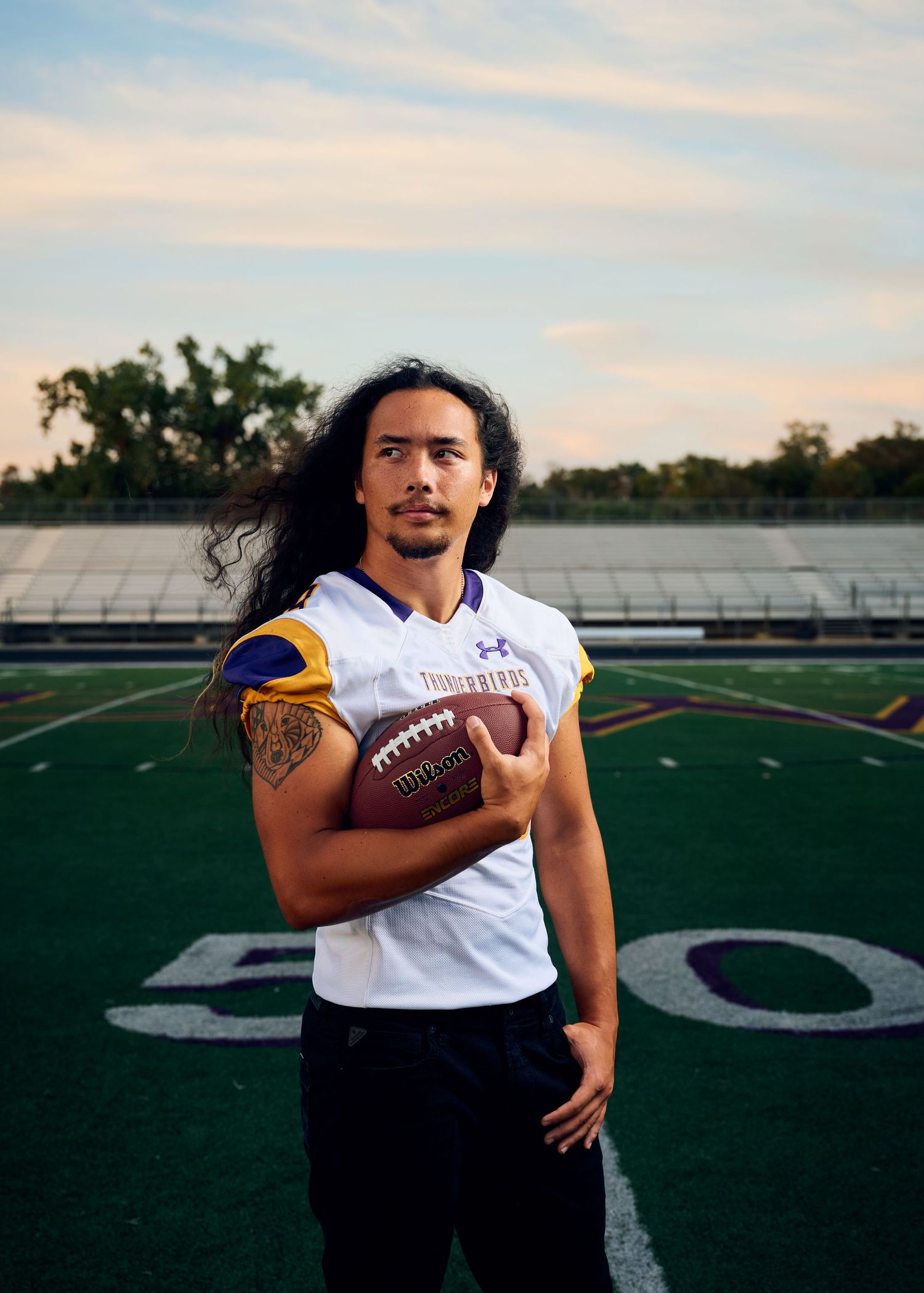 Football player in white jersey stands on field at sunset holding ball with visible tattoo on arm.