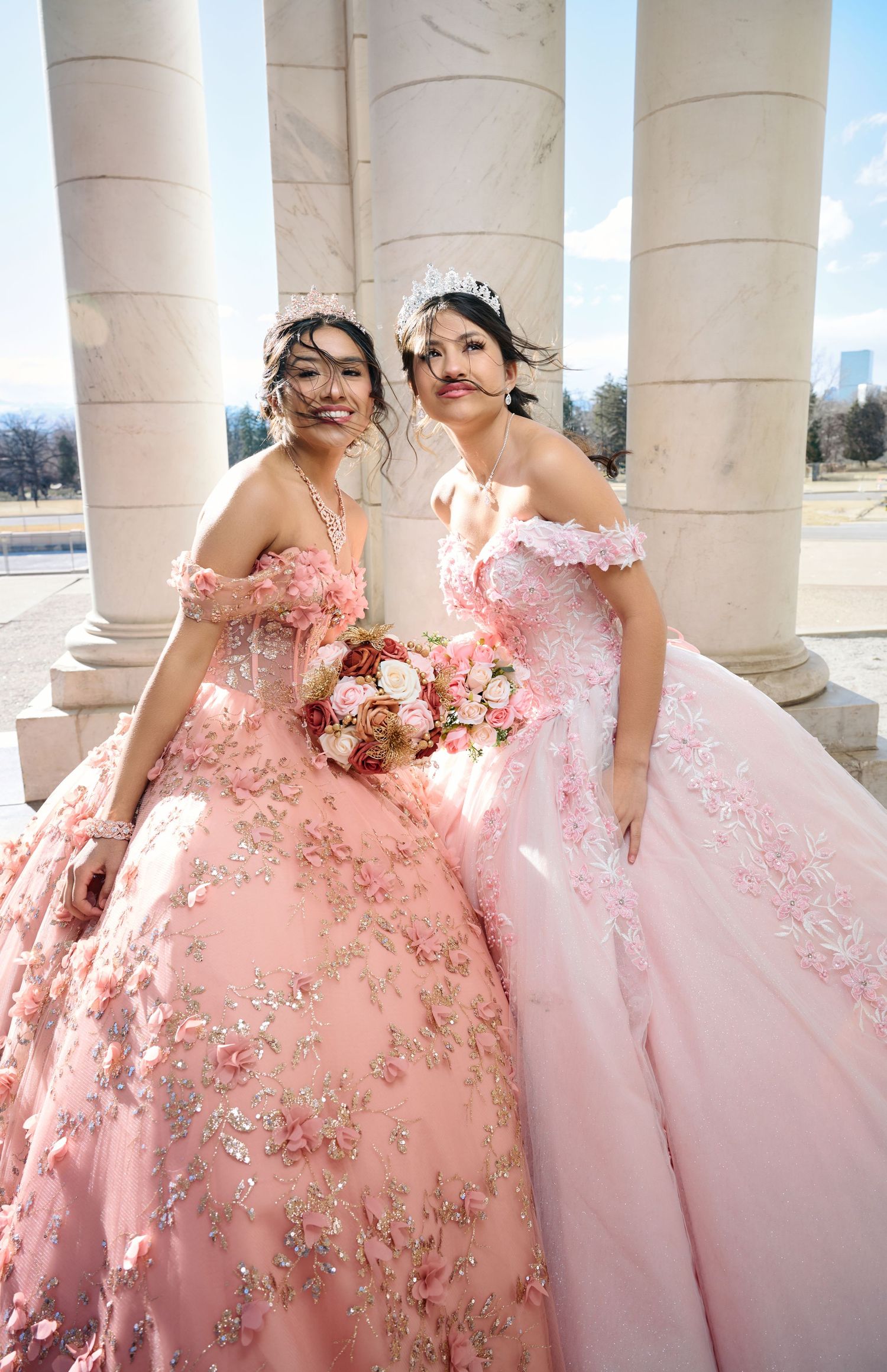 Two women in pink floral quinceañera dresses pose elegantly against white pillars outdoors.