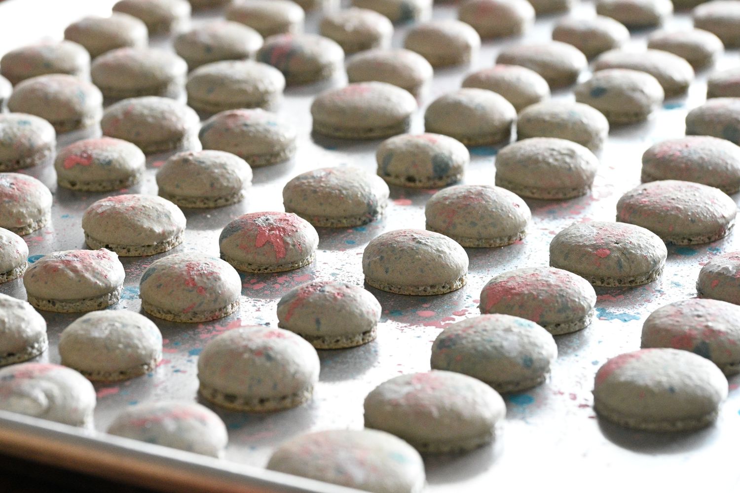 Rows of freshly baked gray macarons with pink centers arranged neatly on a white baking sheet.