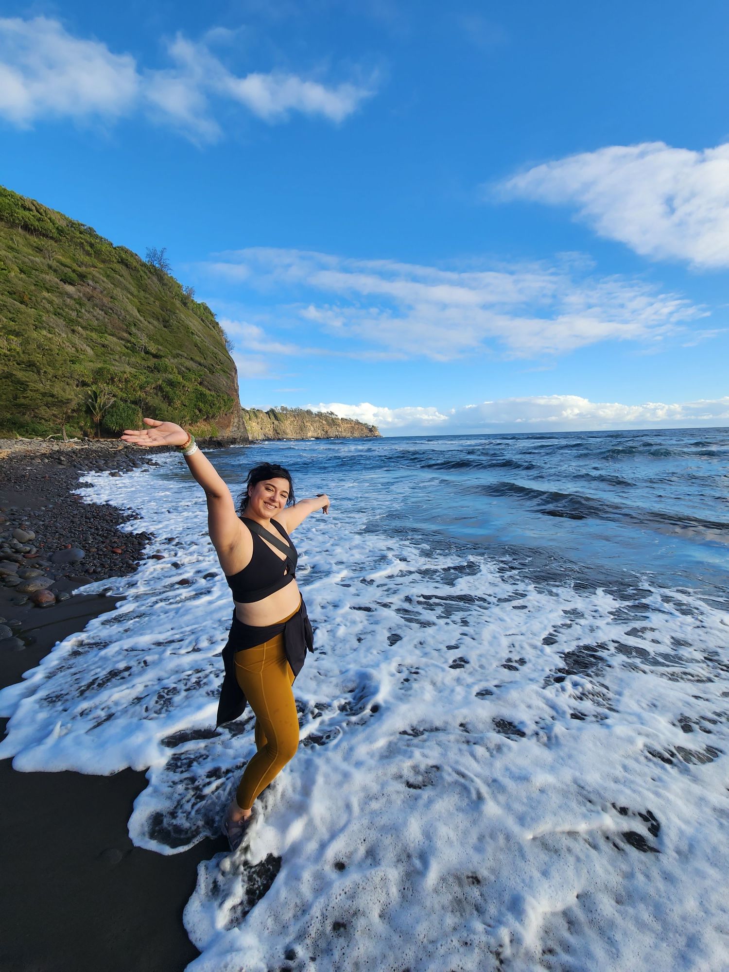 An energetic pose captured on a black sand beach with ocean waves and cliffs in background.