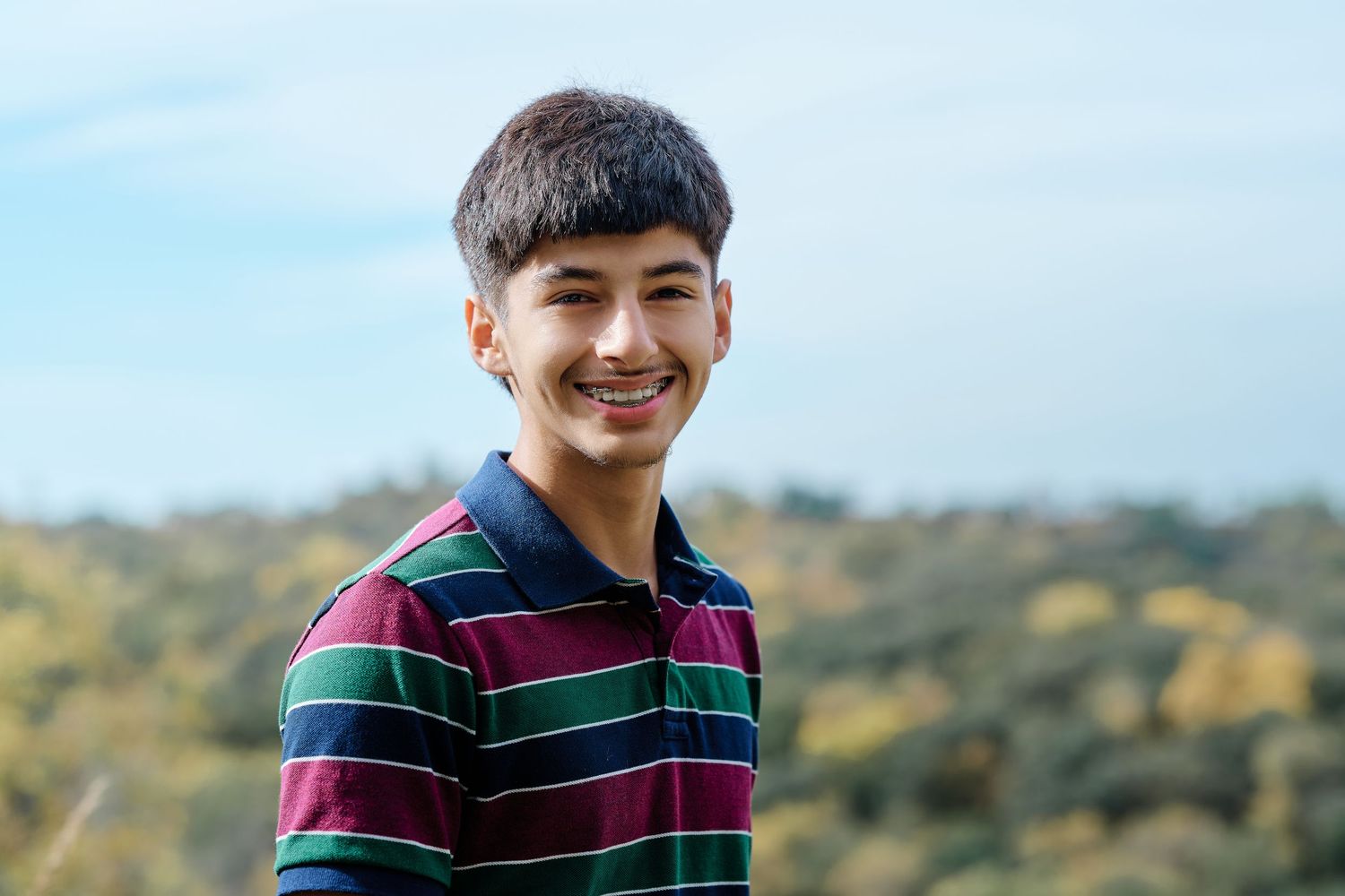 A person wearing a navy and green striped polo shirt smiles warmly while standing outdoors against a blurred nature background.