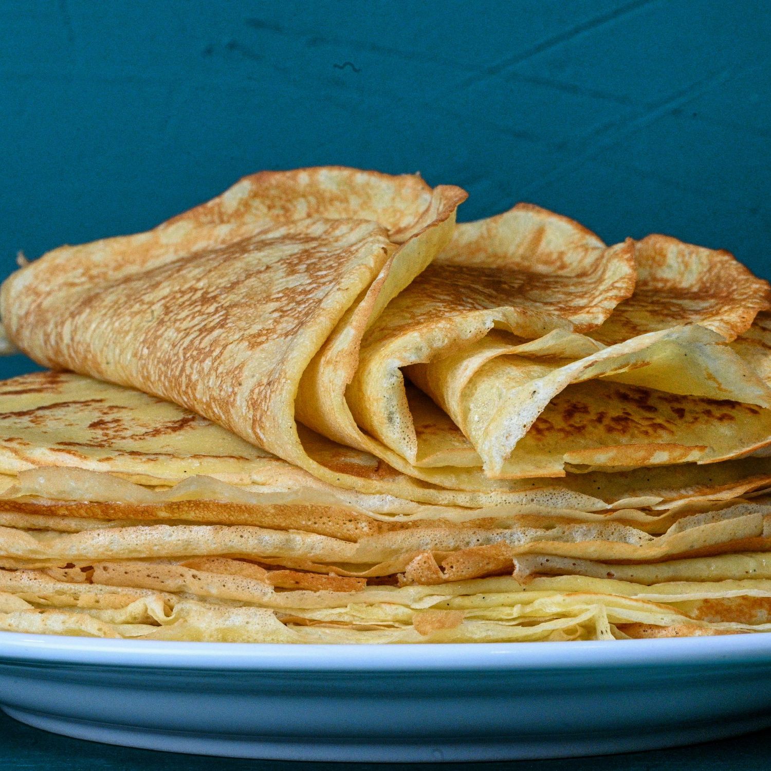 A stack of thin, golden French crepes folded and layered on a white plate against a deep teal background.
