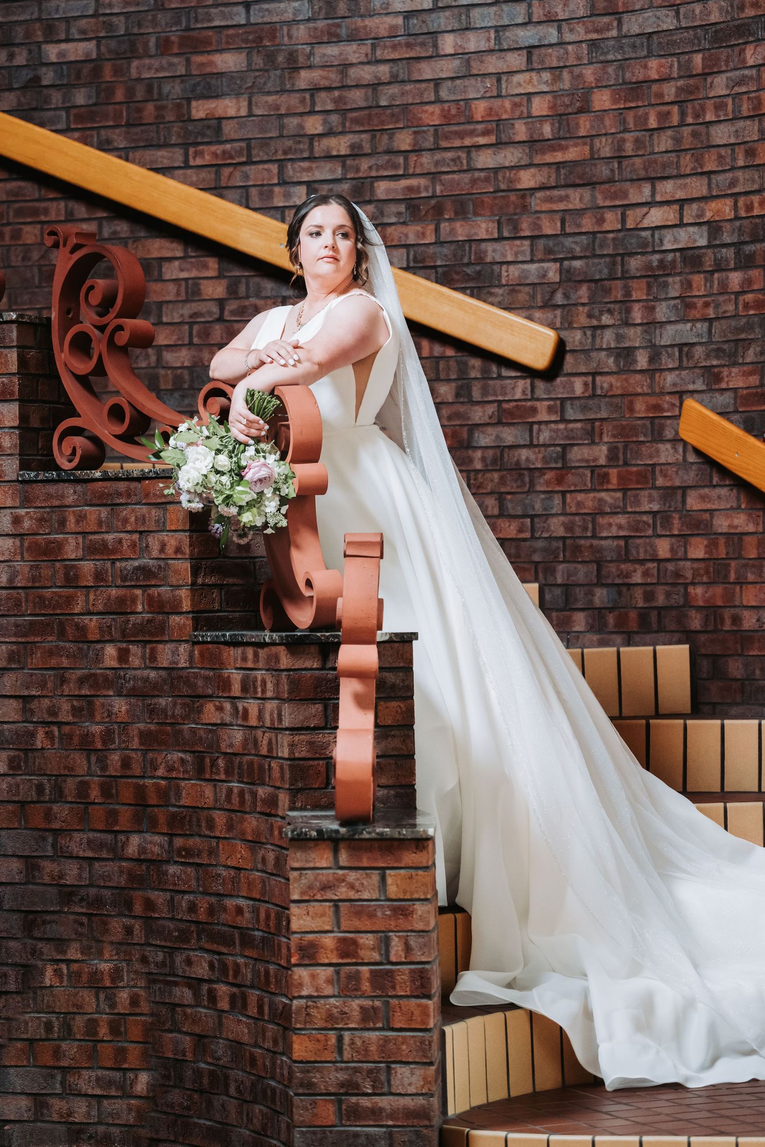 A bride in a flowing white dress and veil poses on rustic brick staircase with ornate railing and white floral bouquet.