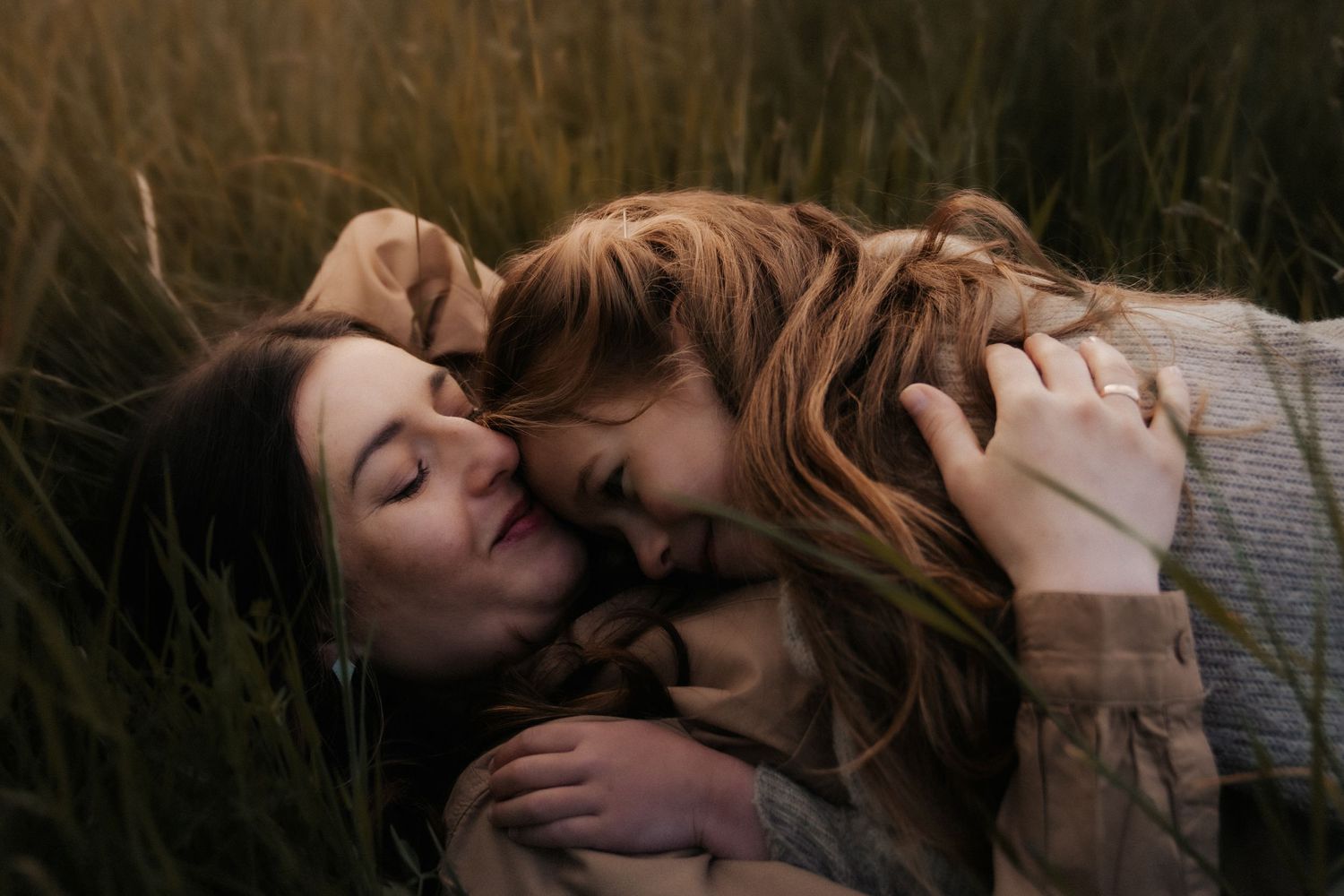 Two people share a tender kiss while laying in tall grass during golden hour lighting.