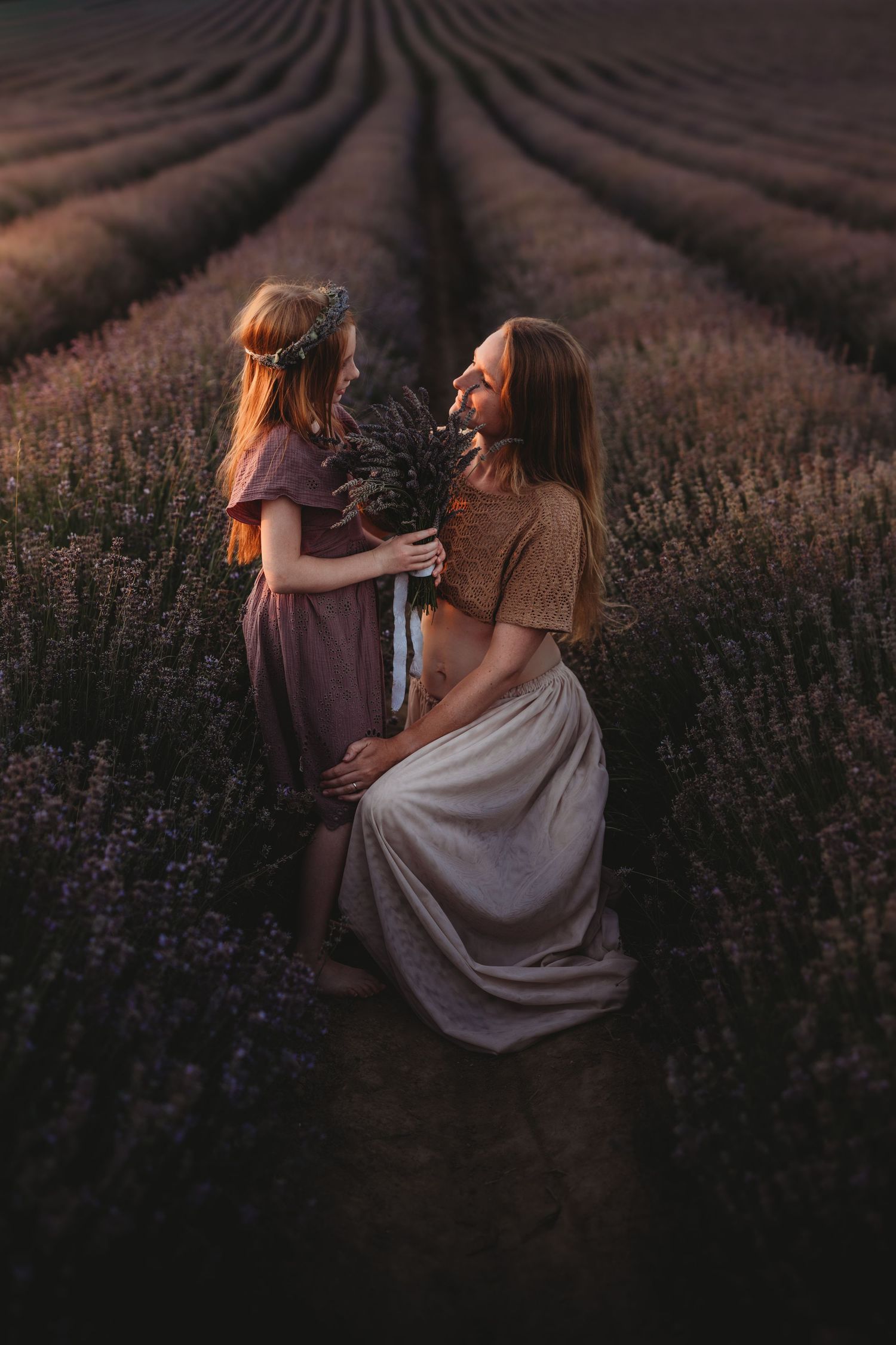 A mother and daughter share a tender moment in a blooming lavender field, holding a bouquet of purple flowers.