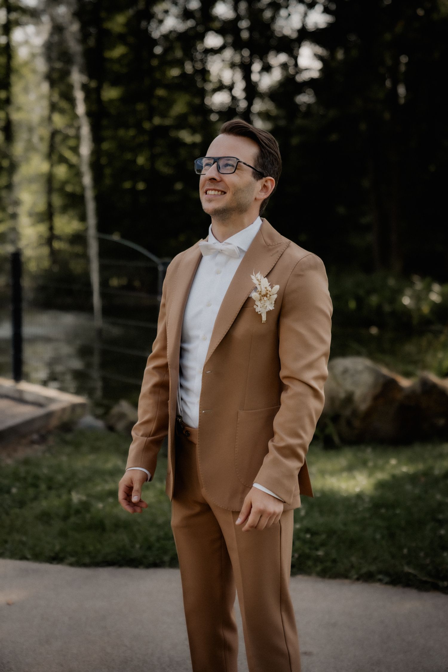 Groom in tan suit and bow tie stands on garden path.