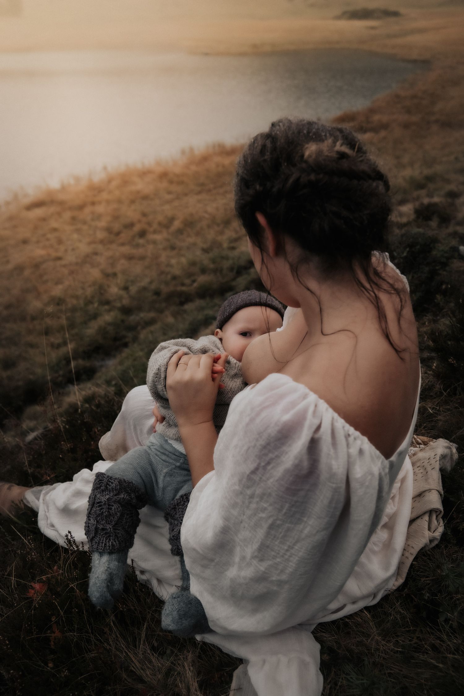 Figure in white dress cradles infant on grassy hillside at sunset.