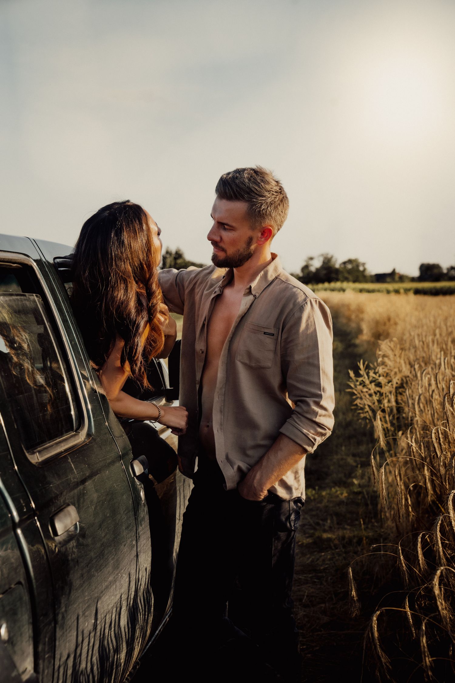 Couple sharing an intimate moment next to black vehicle in wheat field at sunset.