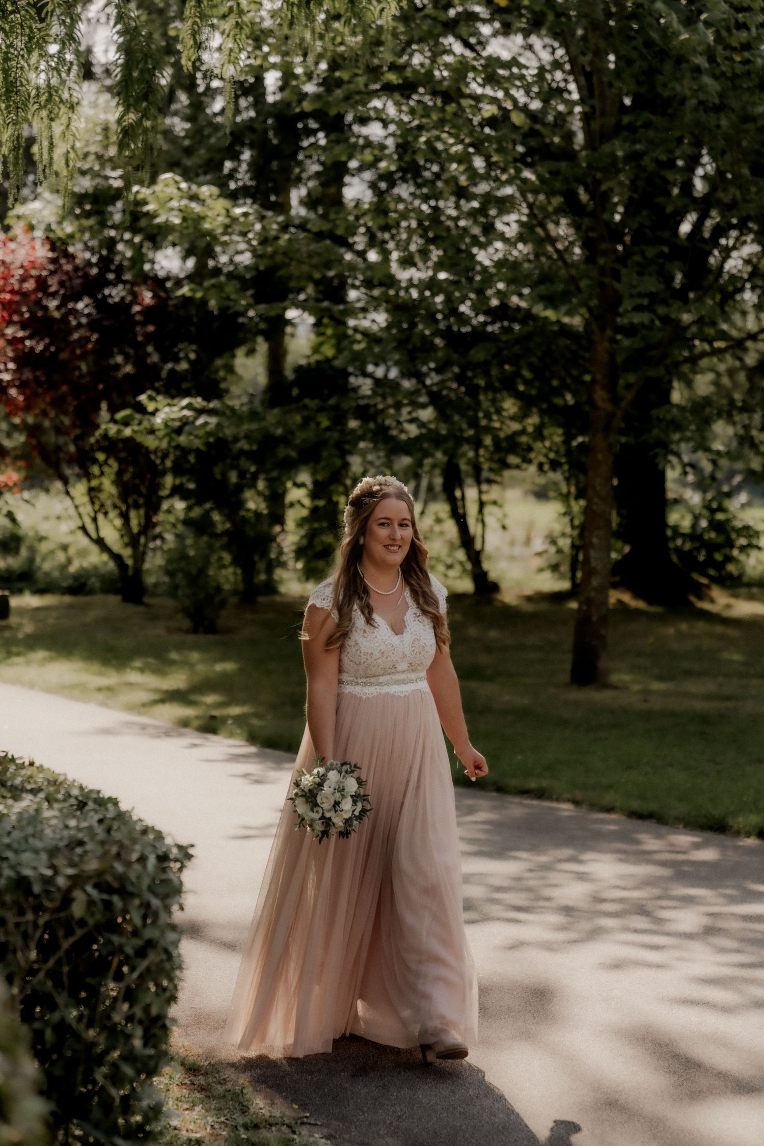 Bride in flowing blush gown holds bouquet while walking garden path.