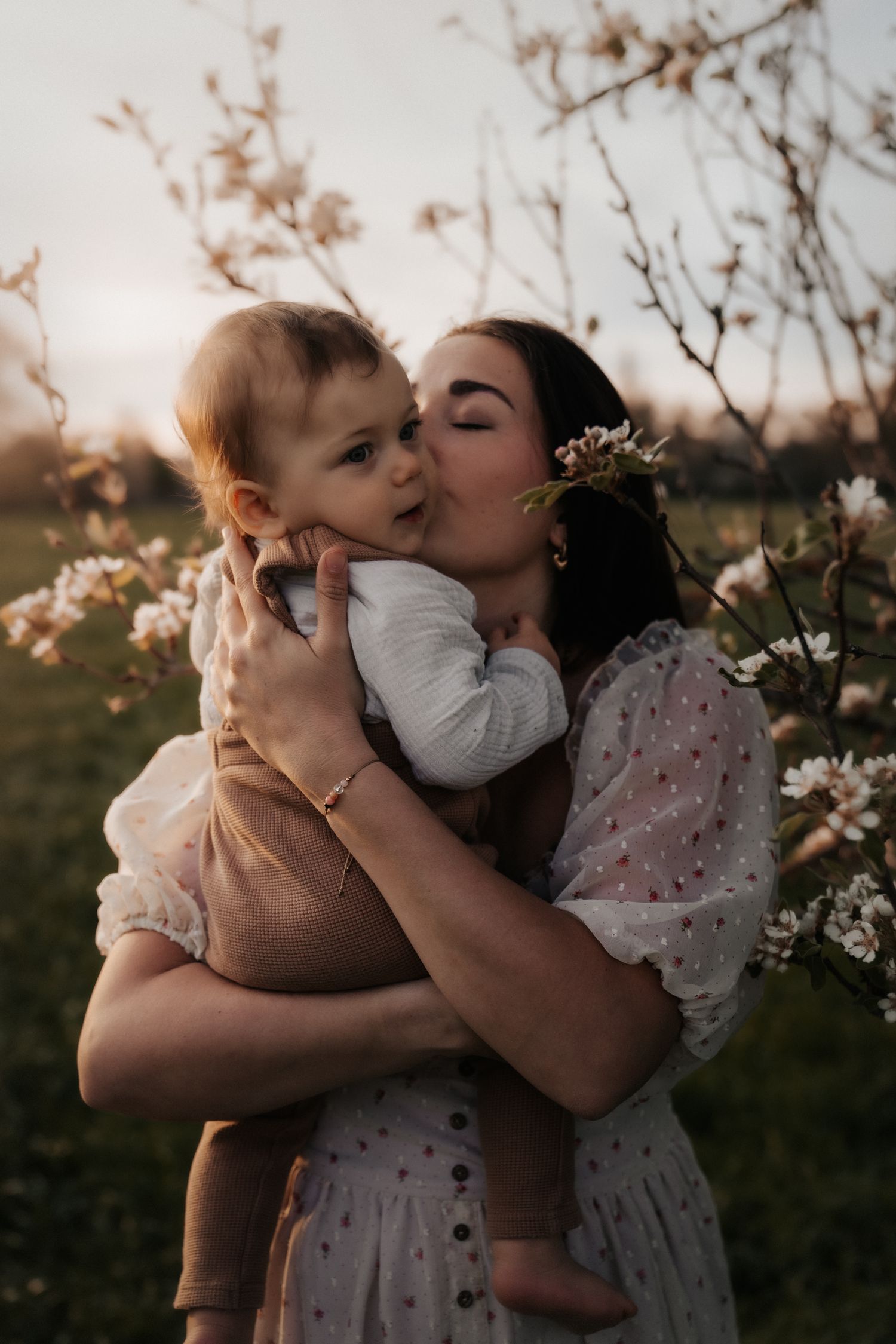 A mother tenderly kisses her baby outdoors near blooming spring trees at golden hour.
