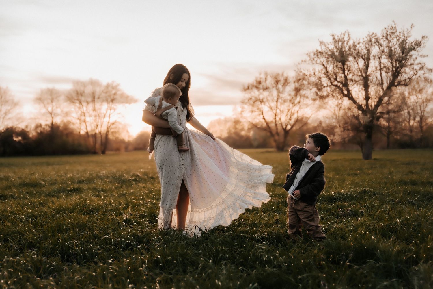 A mother in a white lace dress holds a baby while a young boy looks up at her during a golden sunset in a field.