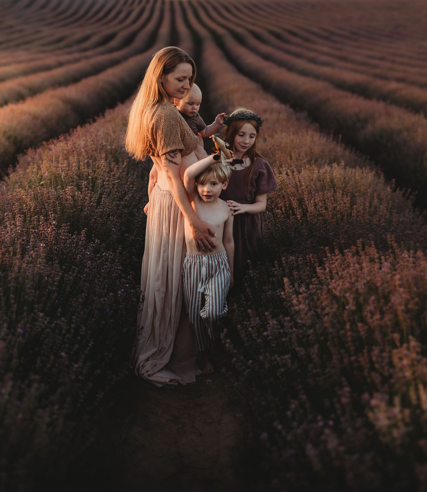 A mother holds three children in a lavender field at golden hour, wearing flowing earth-toned dresses.