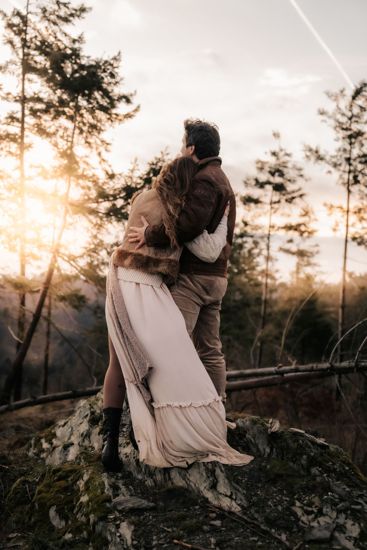 Romantic couple embracing on rocky cliff edge during golden sunset with tall pine trees silhouetted in background.