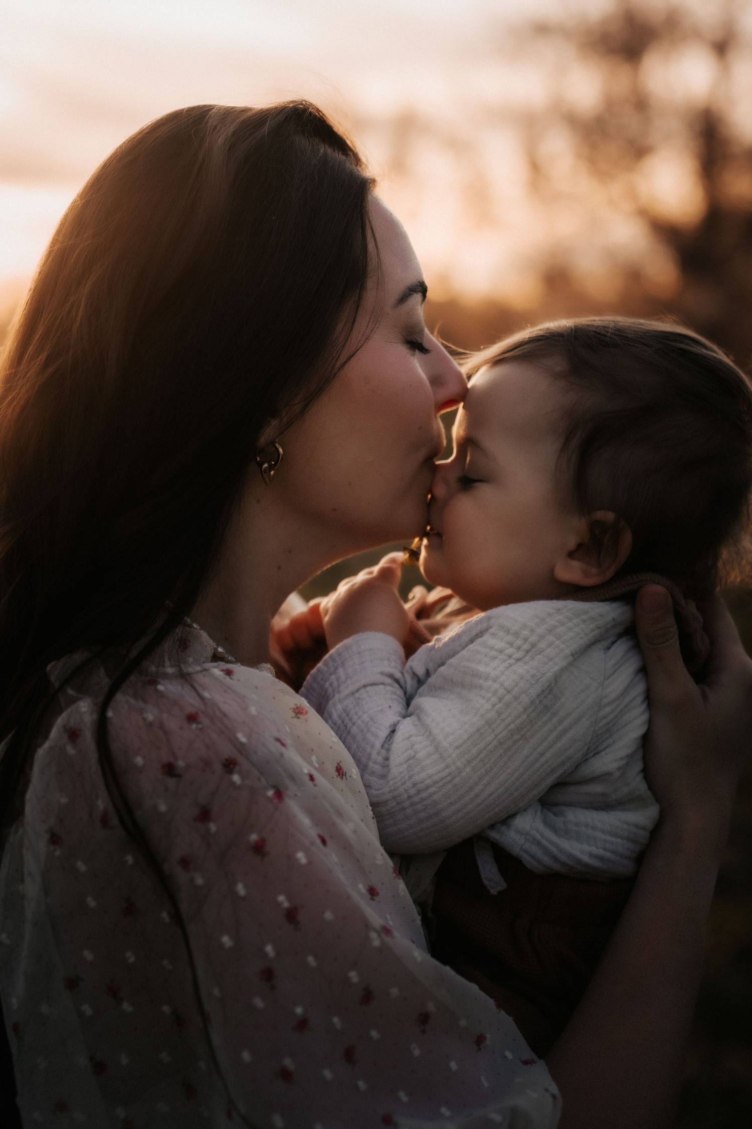 A mother tenderly kisses her baby at golden hour sunset, both bathed in warm light outdoors.