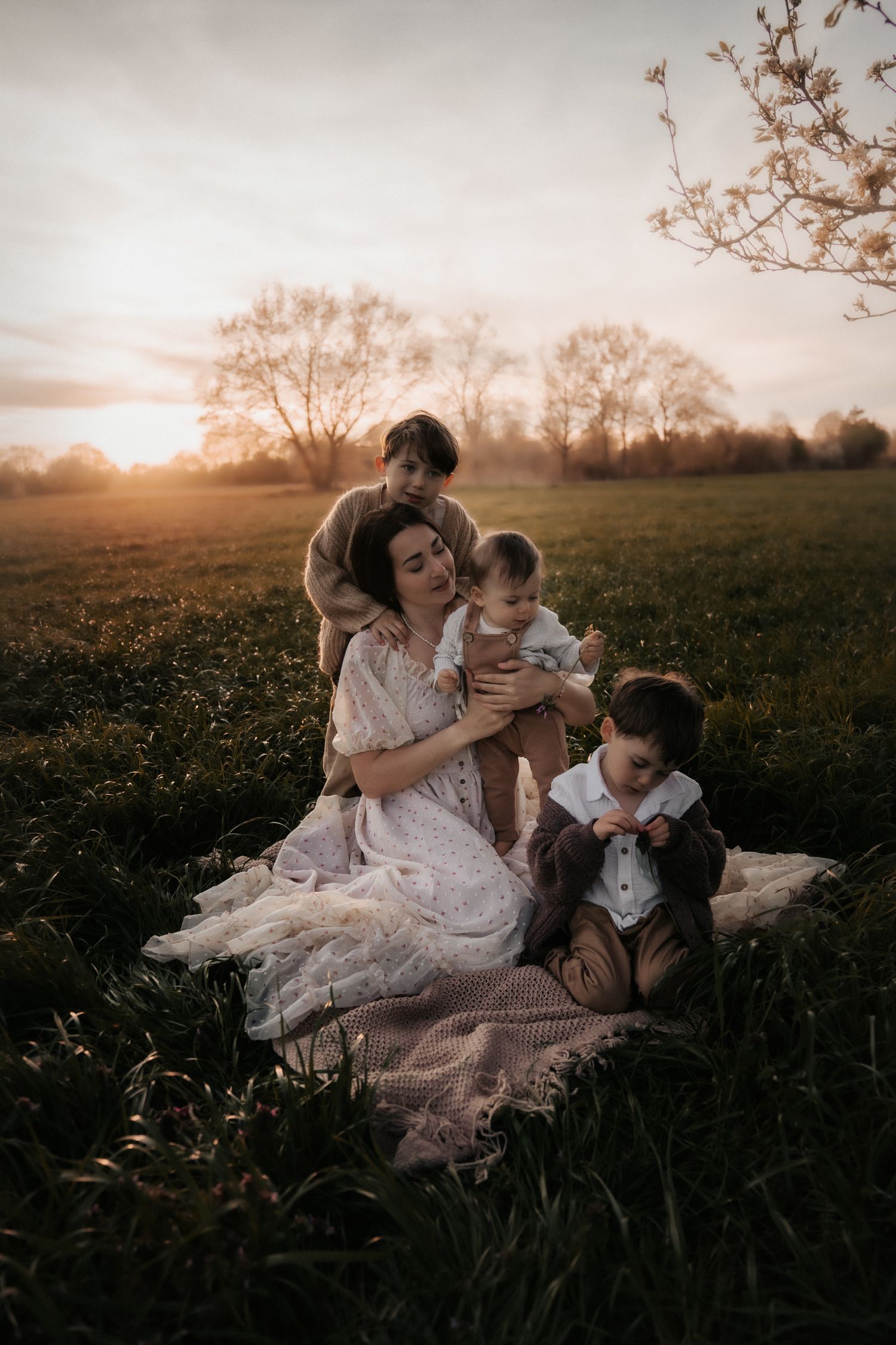 A mother sits on a blanket in a golden field at sunset, tenderly holding her baby surrounded by two young children.
