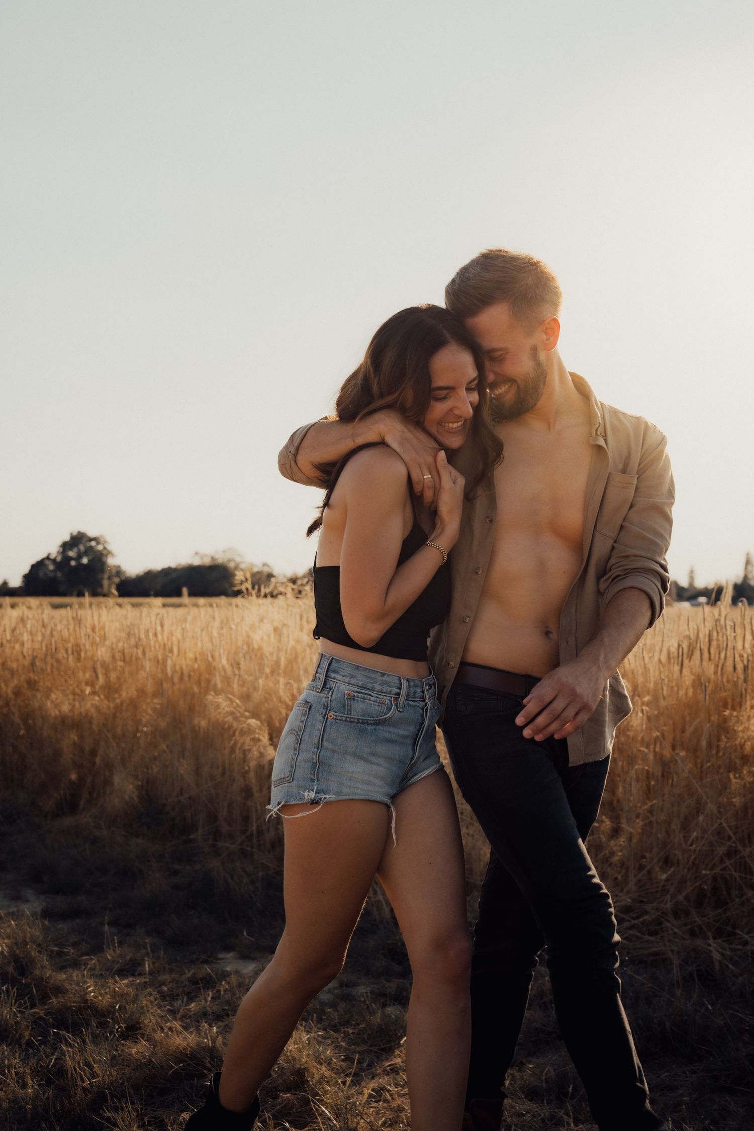 Romantic couple embraces in a golden wheat field at sunset wearing casual summer outfits.