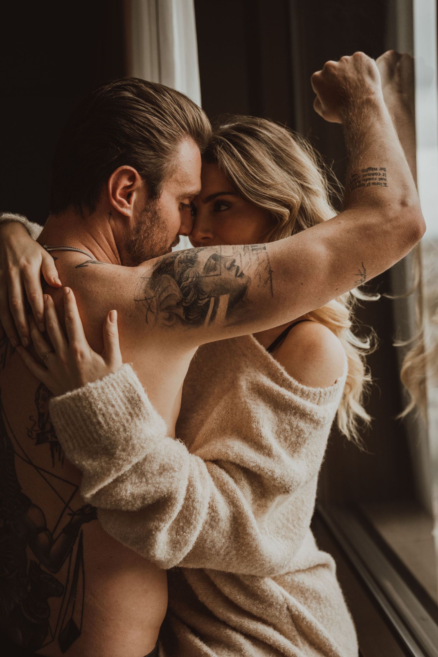 An intimate couple's portrait by a window showing tattoos and a cozy sweater.