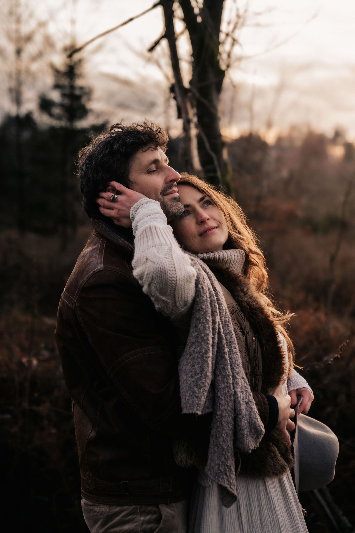 A couple embraces outdoors at sunset in autumn wearing cozy sweaters and scarves in a moody forest setting.
