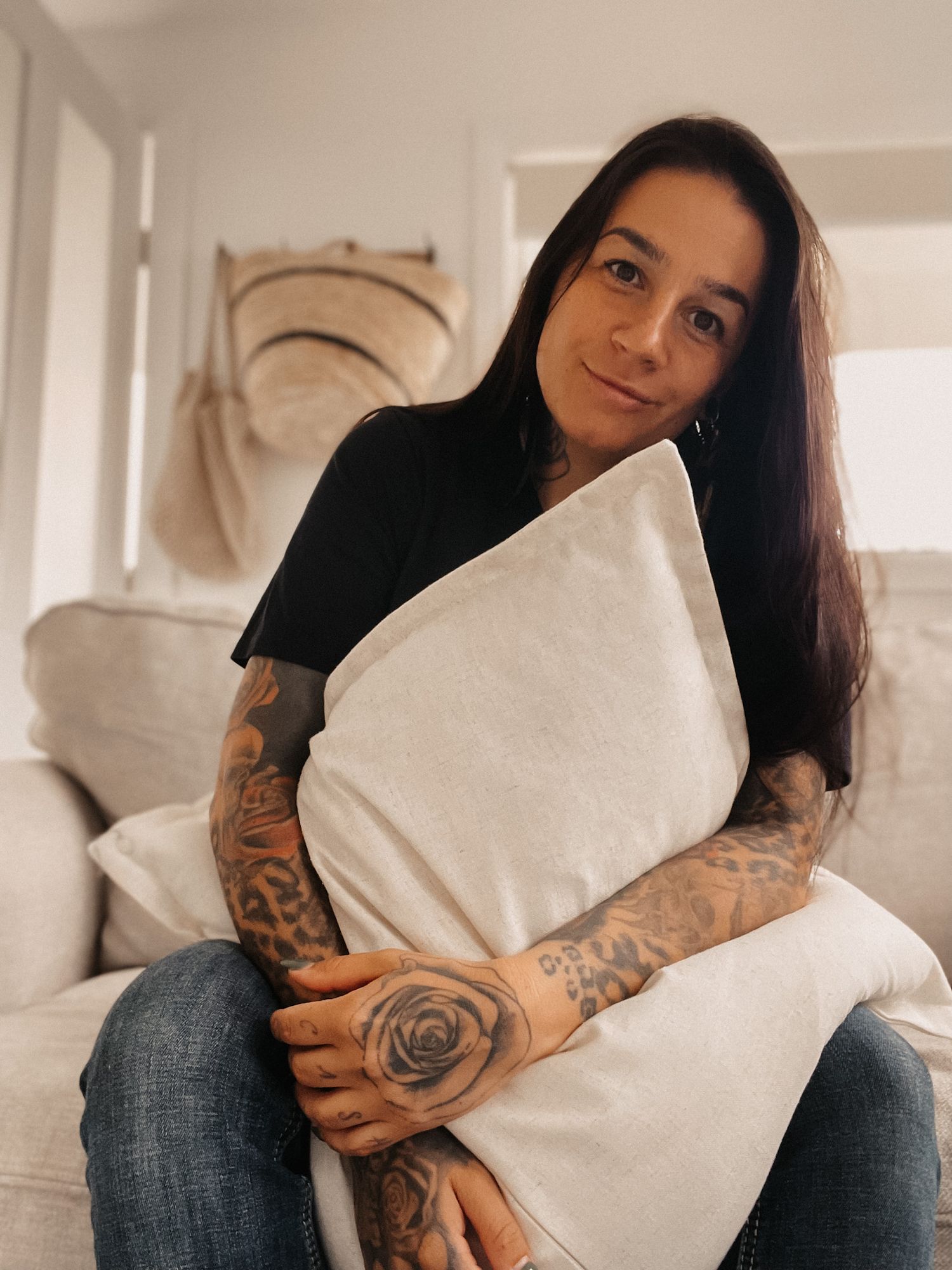 A person with tattoos sitting on a couch holding a white pillow in a bright interior space.