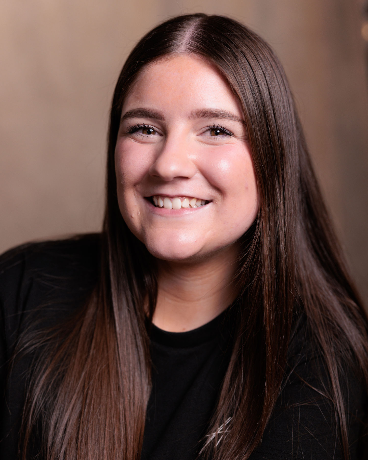 Professional headshot portrait of someone with long dark hair wearing a black top against a neutral background, smiling warmly.
