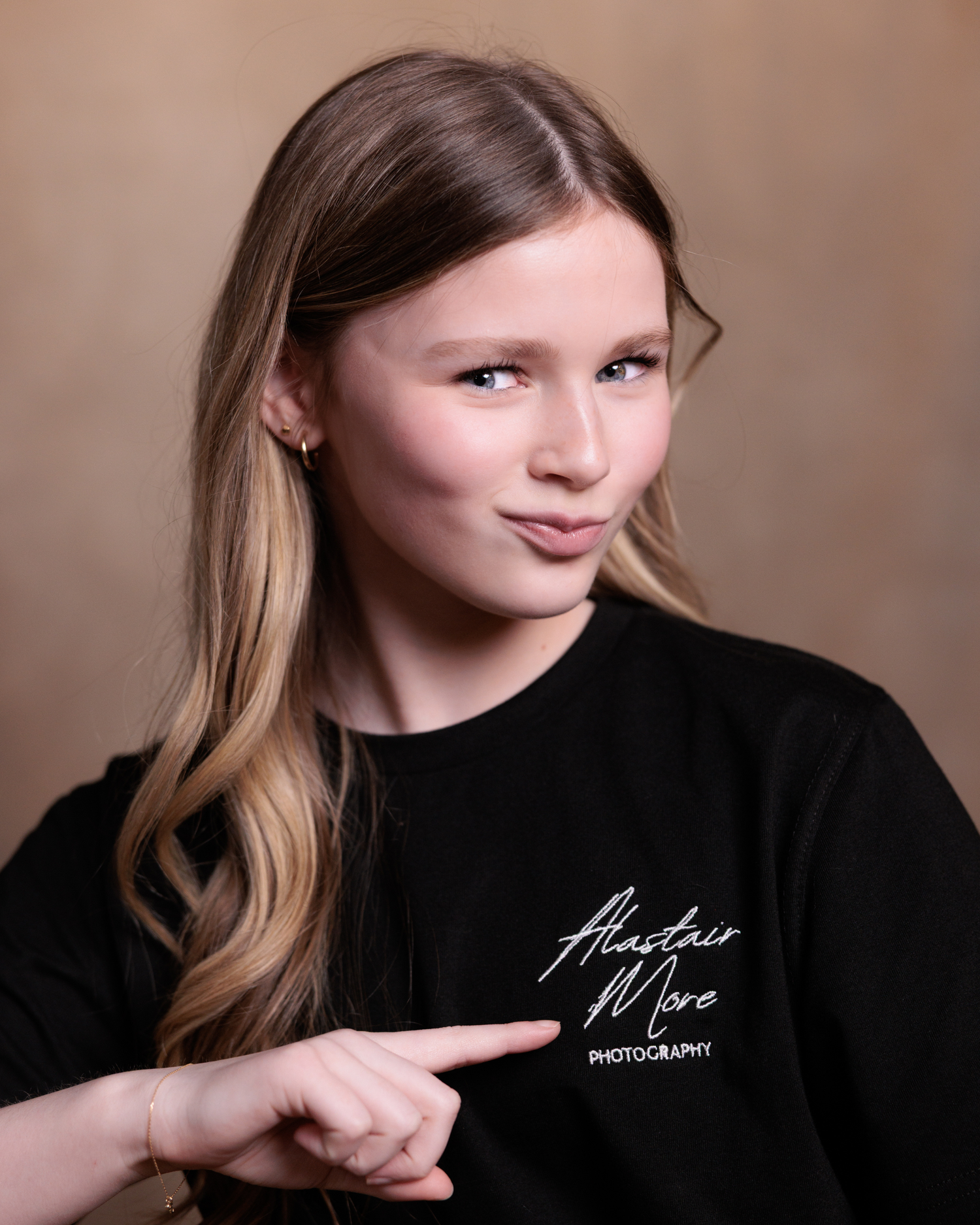 A person in a black sweater with a photography logo points their finger while posing against a neutral background.