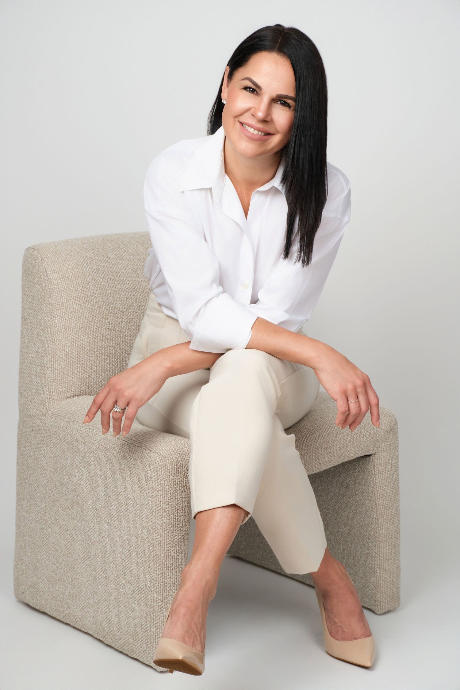 A person in a white shirt and beige pants sits casually on a modern gray upholstered chair in a studio setting.