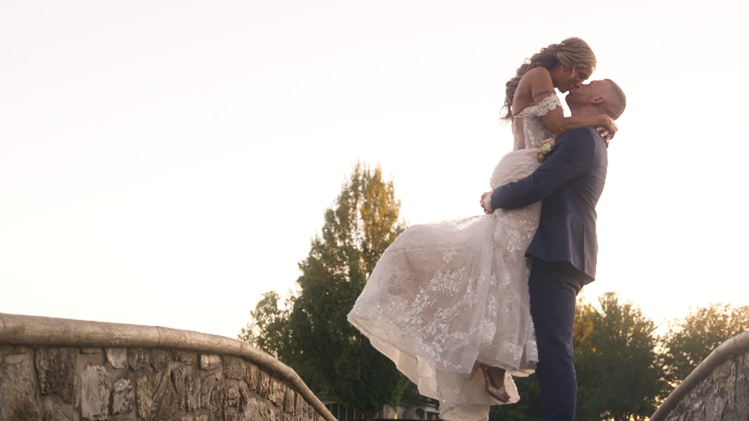 Groom lifts bride for a romantic kiss on a stone bridge against a bright sky.