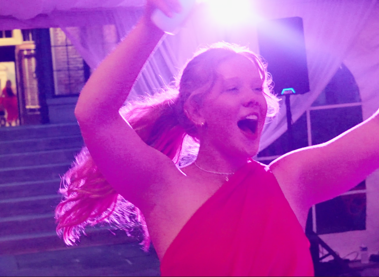 Guest in a pink dress singing and dancing under purple wedding reception lights.