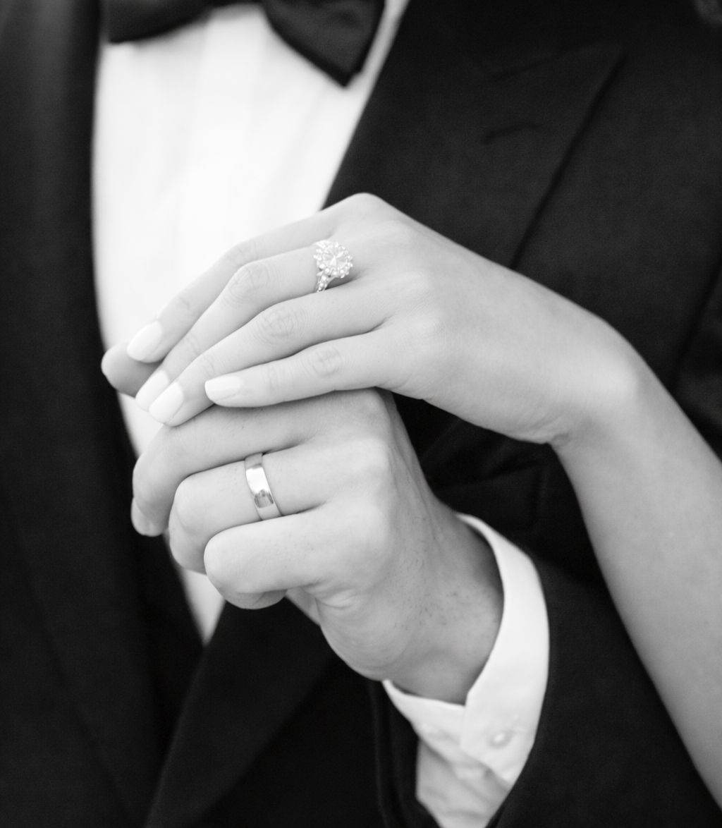 Black and white photo of a bride and groom holding hands, showcasing diamond engagement ring and wedding band.