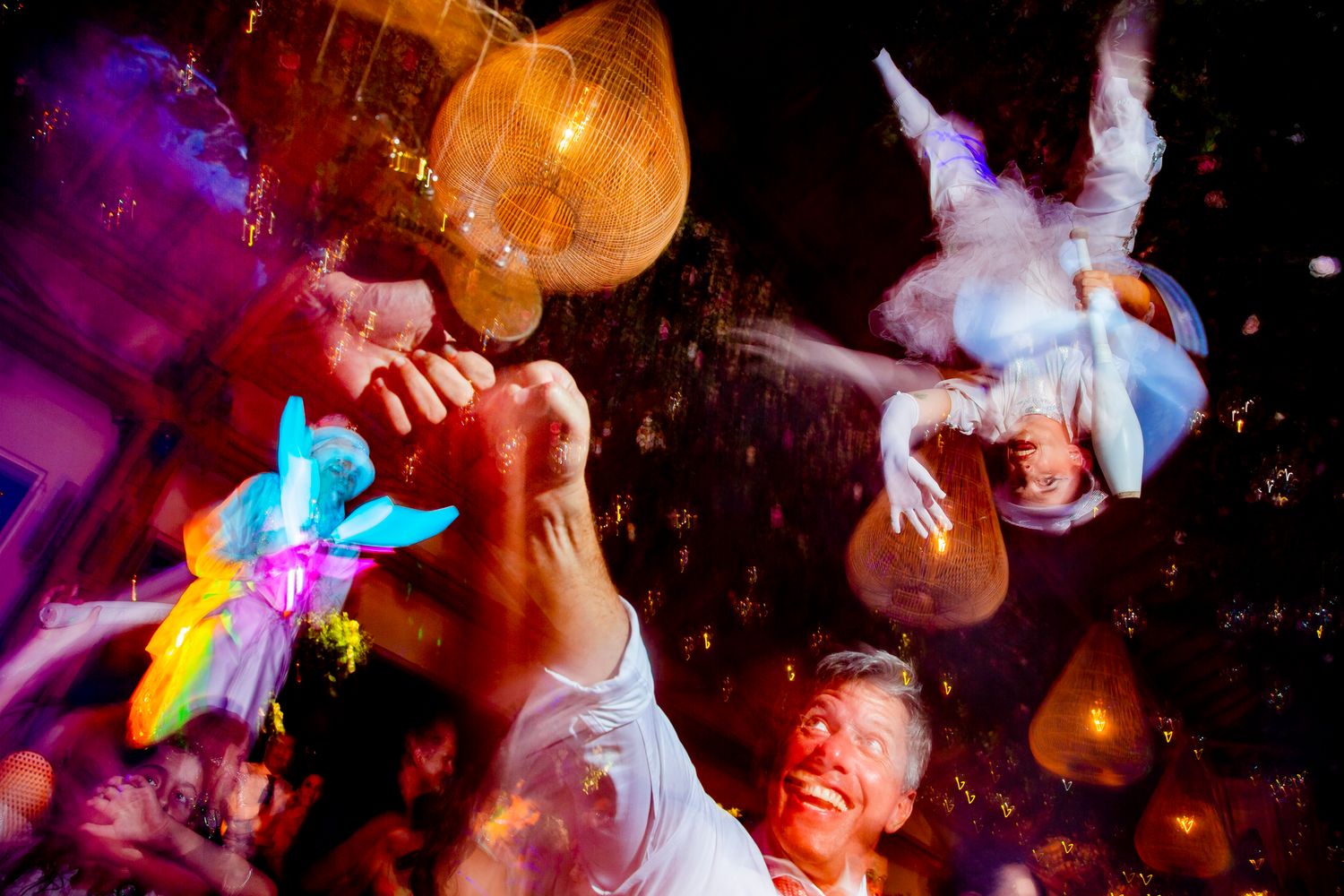 Groom smiling at lively wedding reception party with colorful lights, confetti, and guests celebrating.