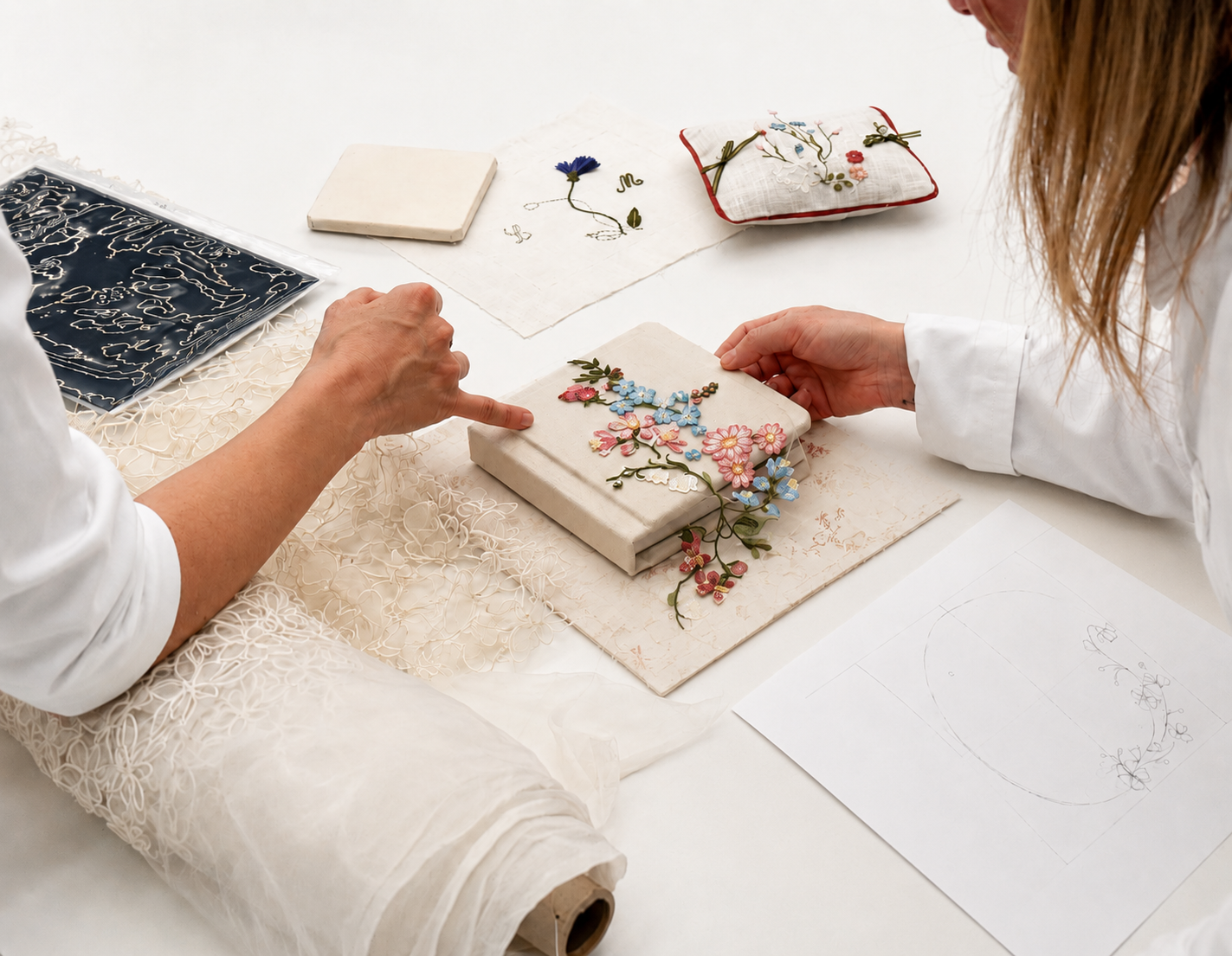 Two people examining delicate floral embroidery on fabric swatches at a white craft table.