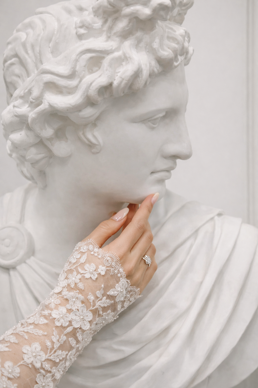 A delicate lace bridal glove positioned against a classical white marble sculpture bust creating an elegant contrast.
