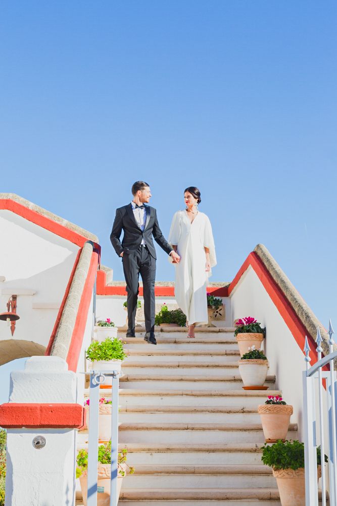 Bride and groom holding hands on white steps with red trim under clear blue sky at Mediterranean wedding venue.