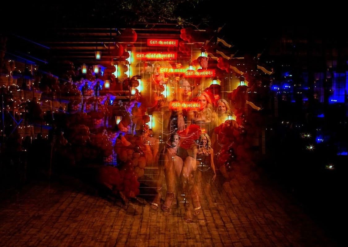 Nighttime street scene with glowing red and blue lights illuminating a crowd on a cobblestone path.