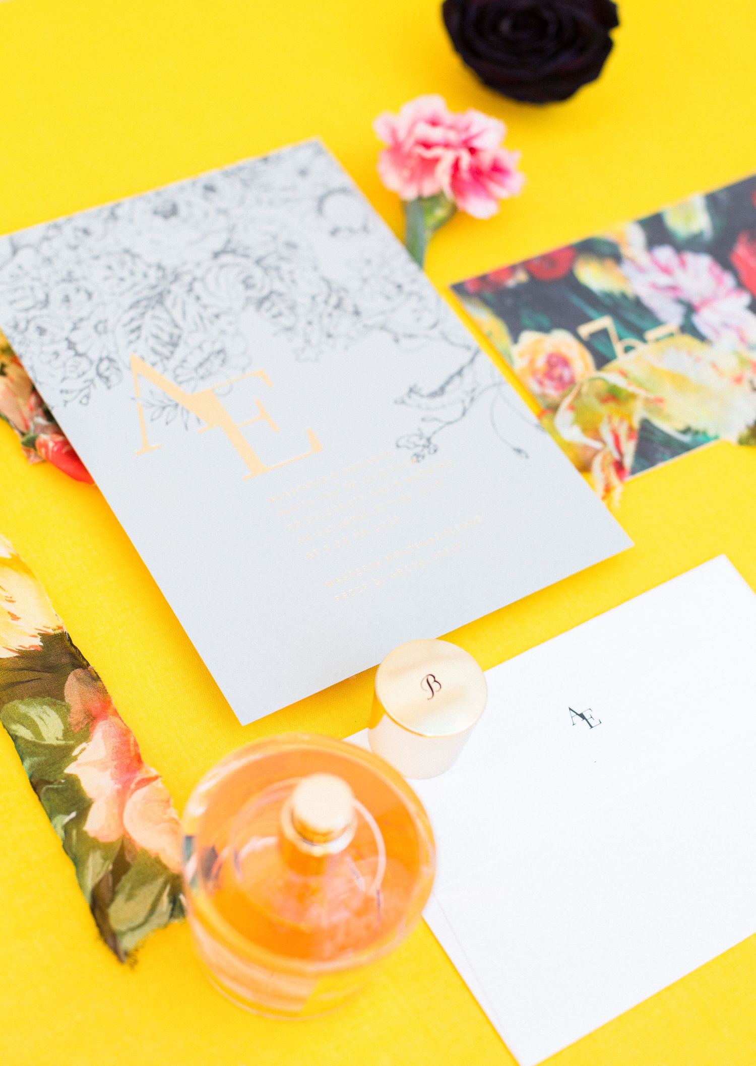 Flat lay of gray floral stationery, pink carnations, floral postcards, and an amber glass jar on a bright yellow background.