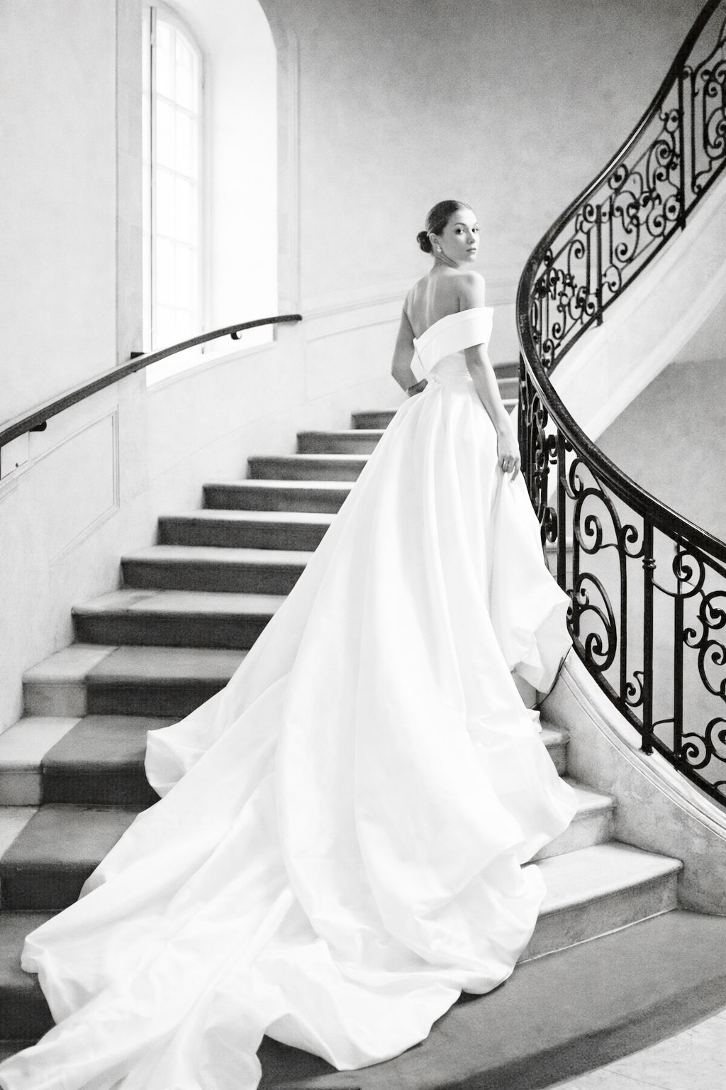 Bride in elegant off-shoulder white gown with long train stands on grand curved staircase with ornate iron railing.