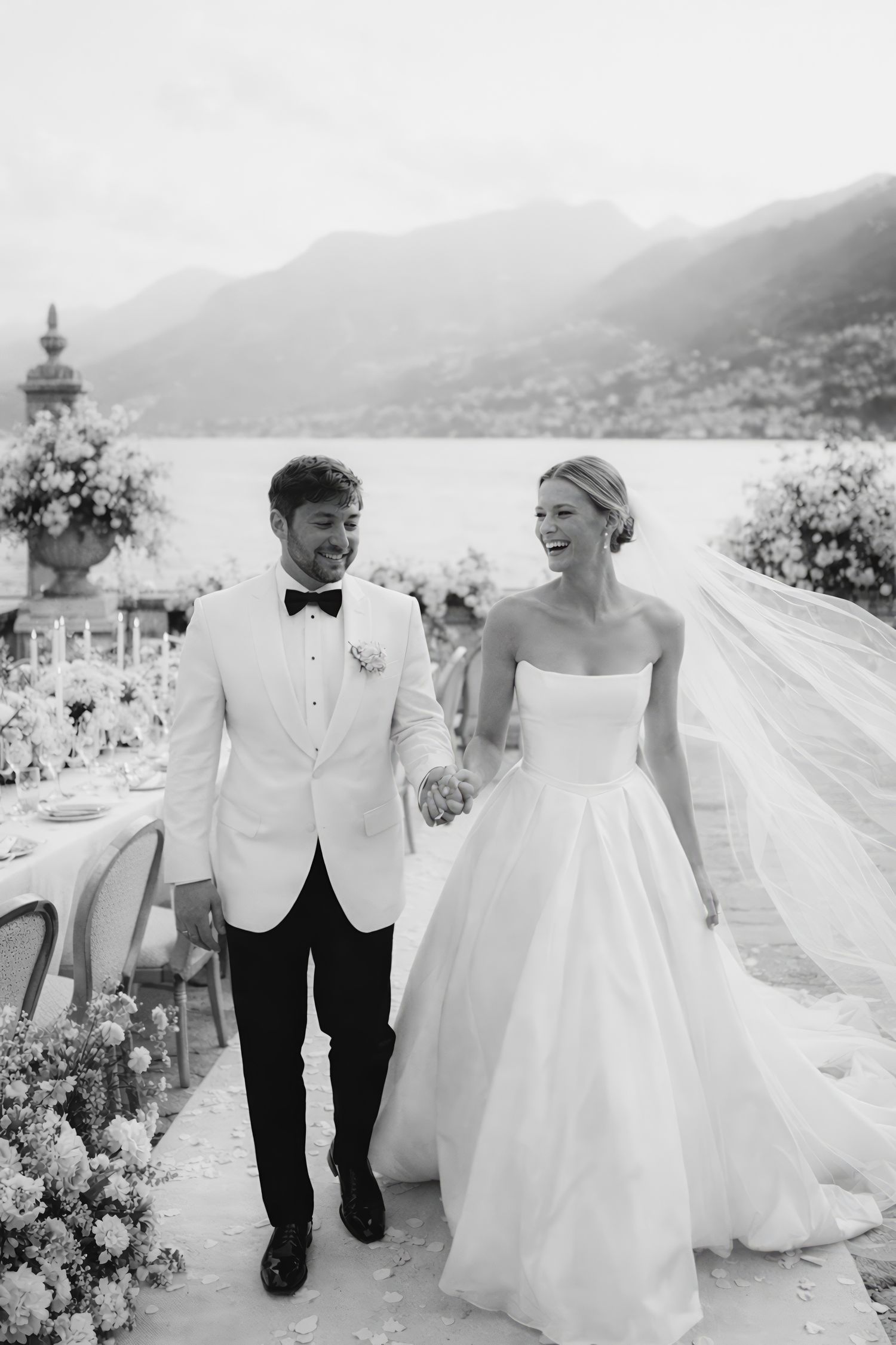 A couple in wedding attire walks together on a scenic mountain path with flowing veil and flowers.