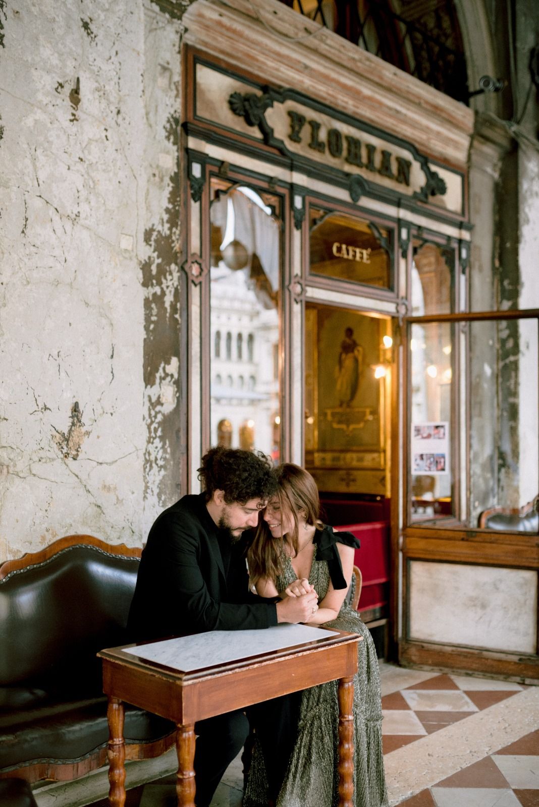 A couple shares an intimate moment at a wooden table inside a vintage cafe with distressed walls and warm lighting.