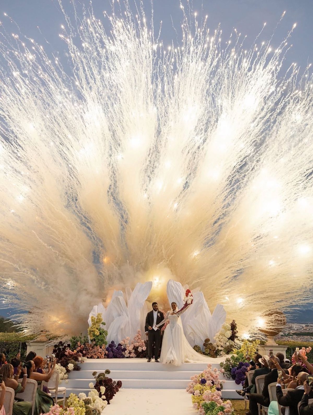 Bride and groom stand on white aisle with stunning fireworks explosion backdrop at elegant outdoor wedding ceremony.