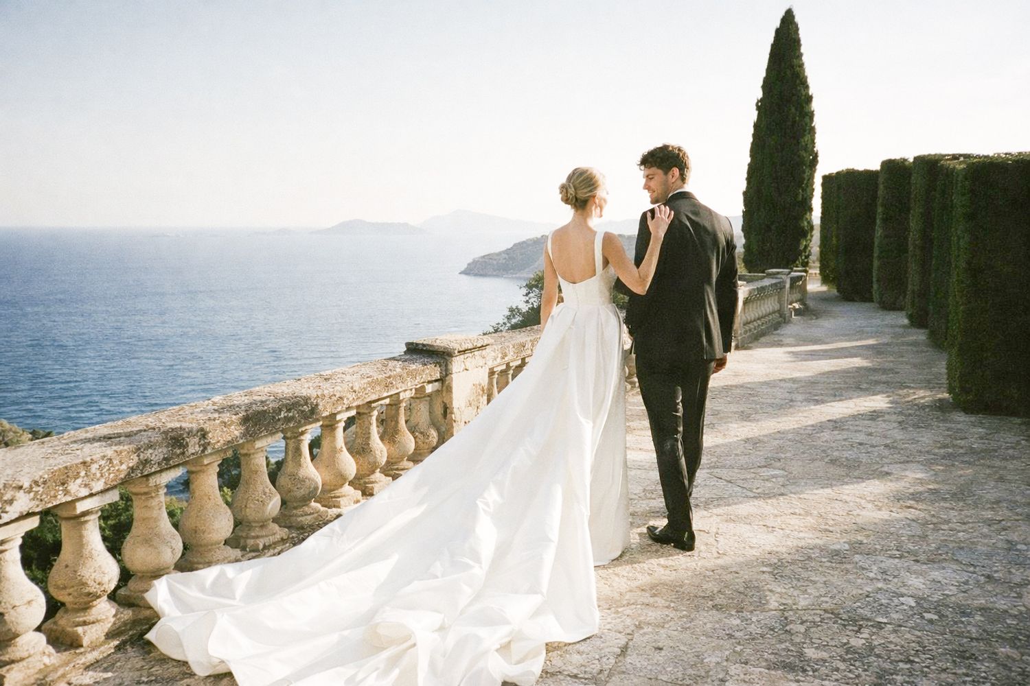 Bride and groom walk along stone balustrade terrace overlooking Mediterranean sea with cypress trees.