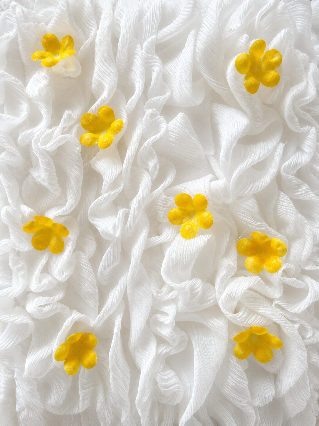 White ruffled fabric texture adorned with small bright yellow flower appliques scattered across the surface.
