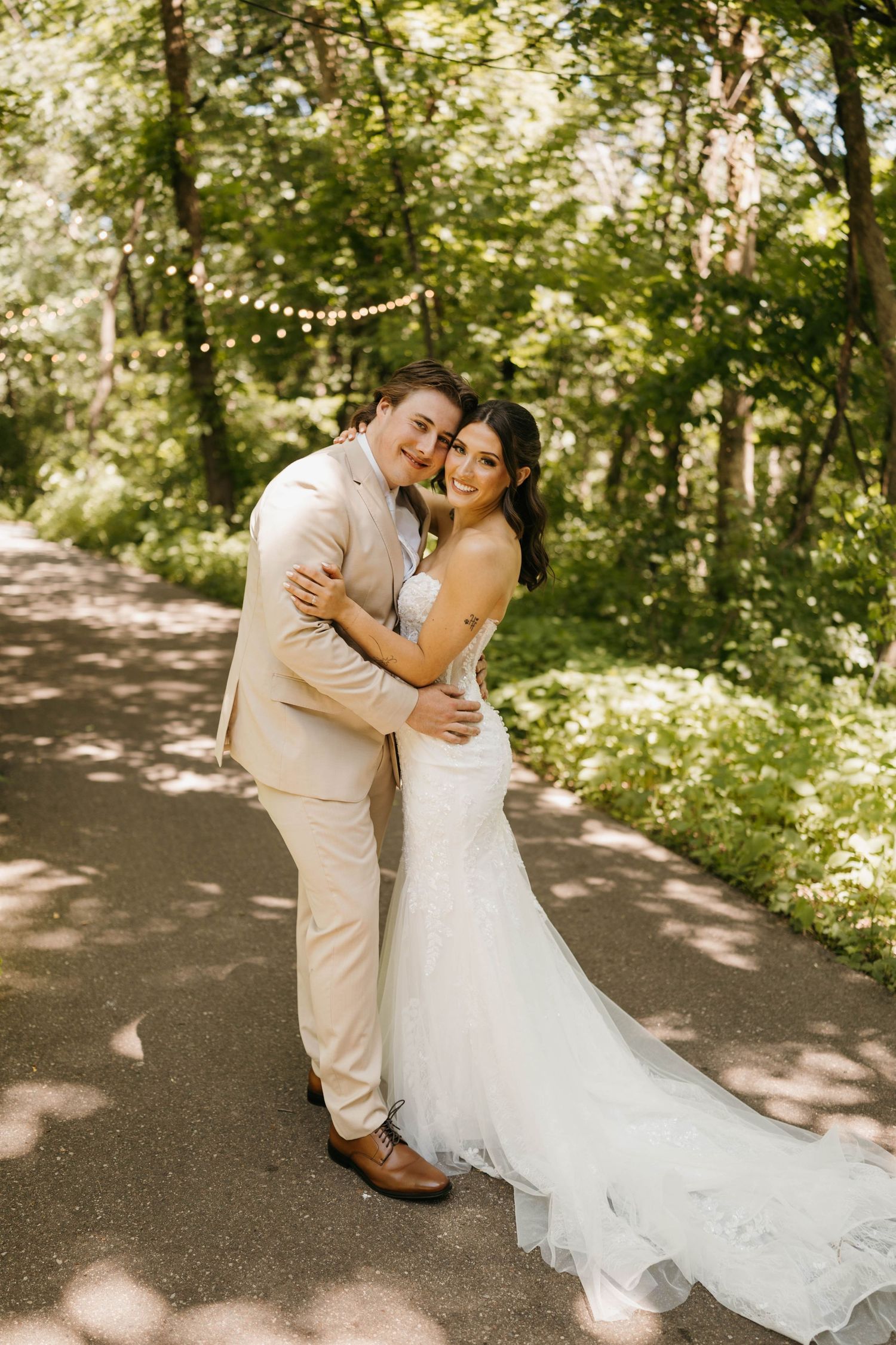Bride and groom embracing on a tree-lined path, wearing a white mermaid gown and beige suit on their wedding day.