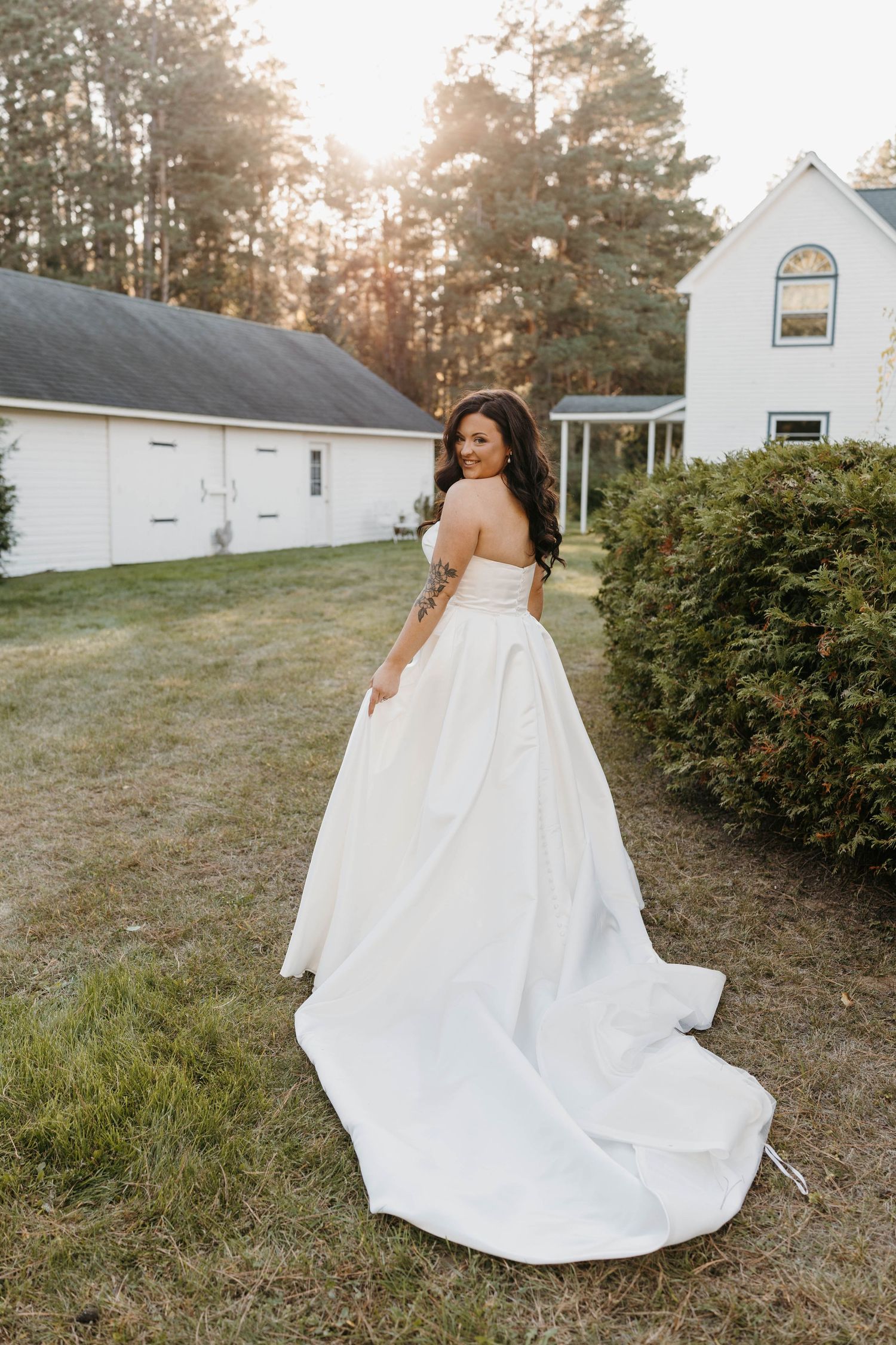 Bride in strapless white ballgown with long train poses outdoors near white farmhouse at golden hour.