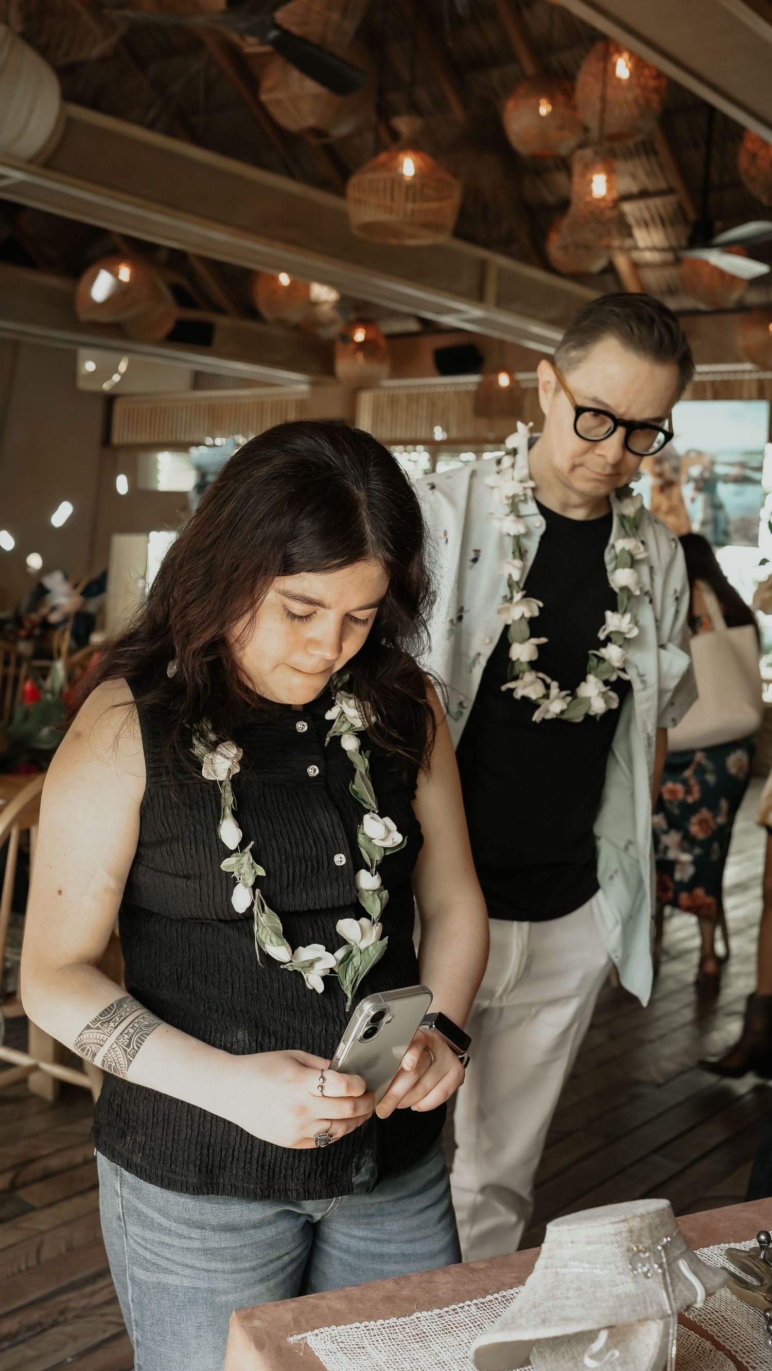 Young woman checking phone and man wearing glasses with floral lei in a warmly lit restaurant.